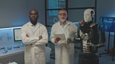 İki farklı robotik mühendisinin orta boy portresi ve insan benzeri robot karanlık laboratuarda kameraya poz veriyor.