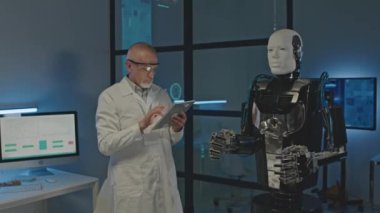 Beyaz laboratuvar önlüklü olgun beyaz mühendisin dijital tablet kullanarak karanlık bilim laboratuarında insansı robotu test ederken orta boy fotoğrafı.