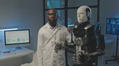 Siyah erkek robotik mühendisinin beyaz laboratuvar önlüklü orta boy portresi karanlık gelecekteki laboratuvarda insansı robotla kameraya poz veriyor.