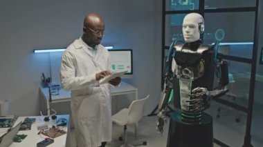 Beyaz laboratuvar önlüklü Afro-Amerikan robotik mühendisinin dijital tablet kullanırken atölyede insan robotu kullanırken orta boy görüntüsü.