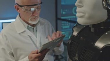 Beyaz laboratuvar önlüklü orta yaşlı beyaz erkek robotik mühendis dijital tablet kullanarak karanlık laboratuardaki robot gibi insanları inceliyor.