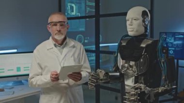 Beyaz laboratuvar önlüklü yetişkin beyaz mühendisin orta boy portresi elinde dijital tablet tutarken karanlık laboratuvarda insansı robotla kameraya poz veriyor.