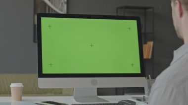 Ofiste masada oturan ve yeşil ekranlı bilgisayar kullanan bir adamın omzunun üzerinden izole şablon ve krom anahtar boş arkaplan ile çalışıyor.