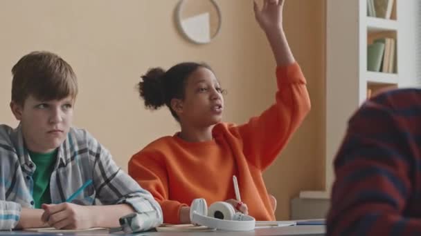 クラスメイトと教室の机に座ってレッスン中に手を挙げるアフリカ系アメリカ人小学生のメディアショット — ストック動画