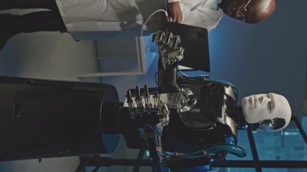 在黑暗车间激活人形机器人时 用笔记本电脑垂直拍摄身穿白色实验室外套的黑人男性工程师 — 图库视频影像