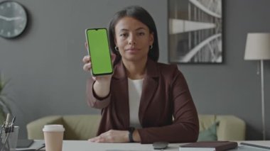 Genç çift ırklı kadının kameraya bakıp minimalist ofiste otururken yeşil ekran ile akıllı telefon sergilediği belden yukarı çekim.