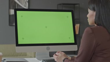 Modern ofisteki beyaz masada oturan, yeşil renkli anahtar model ekran bilgisayara bakan genç, çift ırklı kadının arka plan çekimi.