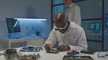 Gece laboratuvarda birlikte çalışırken devre ve anakartların parçalarını inceleyen ve tamir eden iki farklı erkek elektronik mühendisinin orta boy görüntüsü.