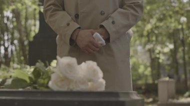 Mezarlıkta güneş ışığıyla aydınlatılan granit mezar taşının üzerindeki beyaz güller ve bulanık arka planda duran deve hendeği ceketli tanınmaz bir adam.
