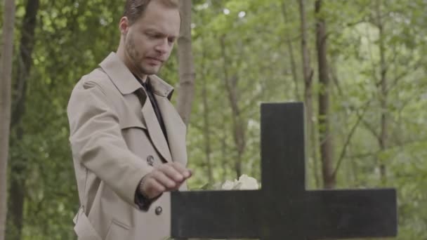 在墓地向墓碑说话和触摸石墨十字架的悲伤男子的中景照片 — 图库视频影像