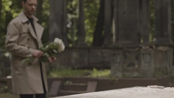 ラクダトレンチコートの白いバラの花束を墓地に持って来る白いトレンチコートのコーカサス悲しい男 — ストック動画