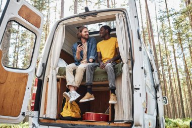 Kamp sırasında ormanda karavanda oturmuş birbirleriyle konuşan iki genç adam.