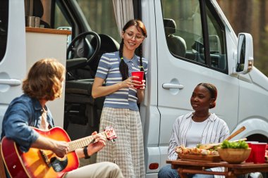 Genç arkadaşlar piknik sırasında arka planda karavanla gitar çalmak için doğada zaman geçiriyorlar.