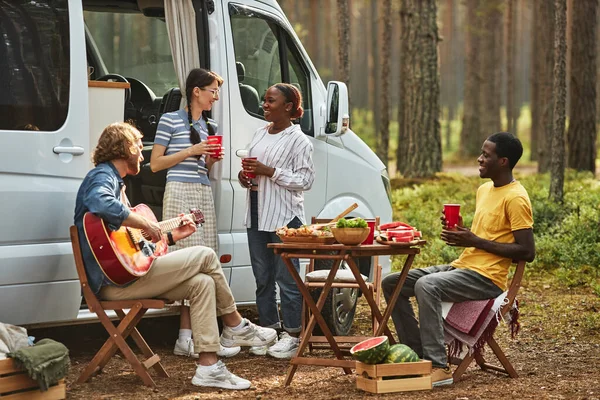 Young people talking to each other and playing guitar while resting in nature during picnic