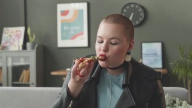 Kafkasyalı, kafası tıraşlı bir kızın gündüz vakti parlak oturma odasındaki koltukta oturmuş lezzetli pizzalar yerken görüntüsü.
