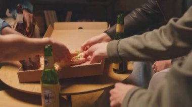 Hafta sonları apartman dairesinde birlikte iyi vakit geçirirken bira içip peynirli pizza yiyen bir grup çok ırklı arkadaşın orta boy fotoğrafı.