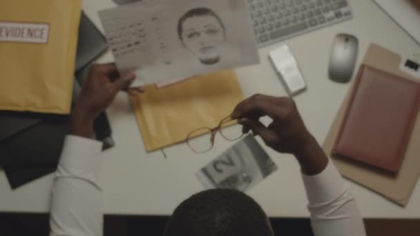 アフリカ系アメリカ人警察探偵のトップビューは オフィスの遅くに刑事事件を調査しながら 印刷された容疑者の同一性を調べている — ストック動画