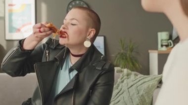 Kafası tıraşlı, siyah deri ceketli beyaz bir kızın ev partisinde arkadaşlarıyla eğlenirken nefis peynirli pizza yediği orta boy bir fotoğraf.