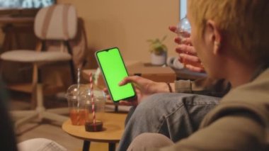 Boyalı sarı saçlı Asyalı çocuğun haftasonu partisinde arkadaşlarıyla takılırken yeşil krom tuşlu akıllı telefon ekranına baktığı omuz üstünden.