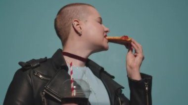 Kendine güvenen, kafası kazınmış beyaz bir kız nefis bir pepperoni pizzasından ısırıyor ve turkuaz stüdyo arka planında duran siyah deri ceketli kameraya bakıyor.
