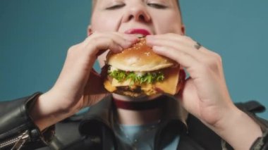 Kafası tıraşlı, kırmızı rujlu, kendine güvenen beyaz bir kızın stüdyosundaki büyük hamburgeri yiyen orta boy fotoğrafı.