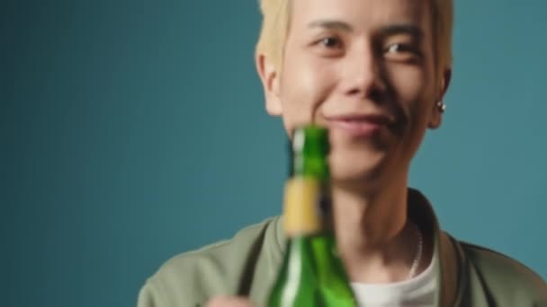 在蓝色摄影棚的背景下 为亚洲金发男子摆姿势 摆上一瓶不含酒精的啤酒 摆出一副相机的样子 — 图库视频影像