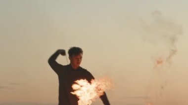 Gün batımında kumsalda ateş gösterisi yaparken yanan nesneleri döndüren ve dans eden kendinden emin bir gencin orta ölçekli fotoğrafı.