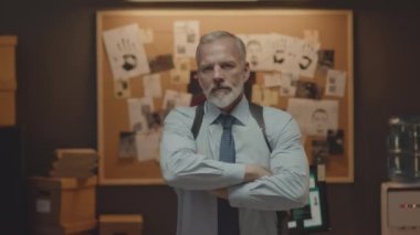 Beyaz saçlı, beyaz saçlı bir polis dedektifinin orta boy portresi. Elleri katlanmış, karanlık ofiste kanıt mantarına karşı poz veriyor.