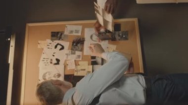 İki çok ırklı FBI ajanının, gazete kupürleri, olay yeri fotoğrafları ve diğer kanıtları, gece ofiste çalışırken, soruşturma mantar panosuna yapıştırdığı dikey bir fotoğraf.