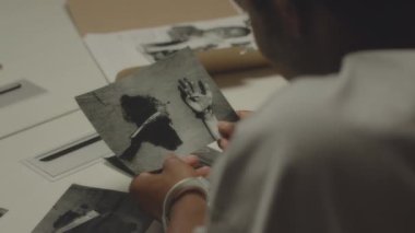 Sorgu odasındaki siyah beyaz olay yeri fotoğraflarına bakan kelepçeli Afrikalı Amerikalı şüphelinin omzunun üstünden.