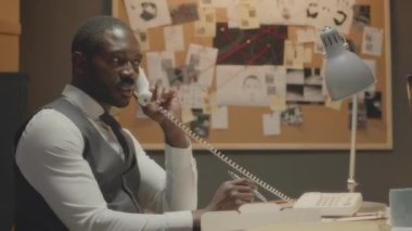 Profesyonel Afro-Amerikan erkek dedektifin ofis masasında oturduğu ve suç davasını araştırırken sabit hattan konuştuğu orta boy bir fotoğraf.