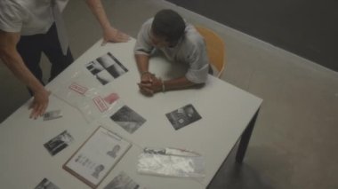 Tutuklu bir adamın, sorgu odasındaki polis tarafından sorgulanırken plastik poşetlerdeki fiziksel kanıtlara ve olay yerindeki siyah-beyaz fotoğraflara baktığı yüksek açılı bir çekim.