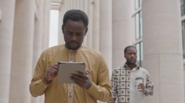 Merkezde gündüz vakti sokakta yürürken dijital tablet kullanan odaklanmış genç Afrikalı Amerikalı adamın orta boy fotoğrafı.