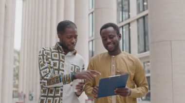 Ilık güneşli bir günde şehir merkezinde yürürken iki Afrikalı Amerikalı genç erkek meslektaşın dosya üzerindeki iş belgelerine bakarken orta ölçekli bir fotoğraf.