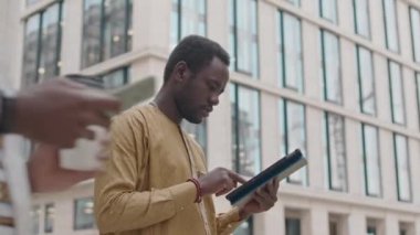 Merkezde, arka planda çağdaş ofis binası varken tablet kullanan odaklanmış genç Afrikalı Amerikalı adamın orta boy fotoğrafı.