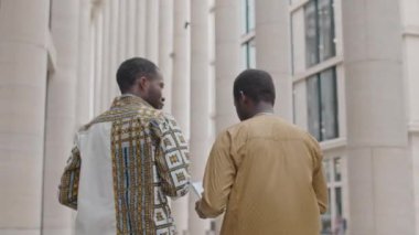 Gündüz vakti şehir merkezindeki ofislerinin önünde yürürken iş belgelerini tartışan iki siyahi erkek iş arkadaşının orta açıdan çekimini takip et.
