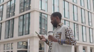Orta boy, şık tişörtlü yakışıklı Afro-Amerikan bir adam akıllı telefondan mesaj atıyor ve şehir merkezinin önünde modern ofis binasına karşı kahve içiyor.