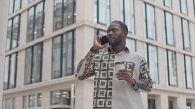 Düşük açılı orta açılı bir fotoğraf, elinde kahve fincanı olan bir genç adam akıllı telefondan konuşuyor ve etrafa bakıyor, arka planda çağdaş ofis binasına karşı duruyor.