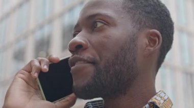 Güleryüzlü Afrikalı Amerikalı genç bir adamın açık havada gündüz vakti telefon konuşması yaptığı orta boy yakın çekim.