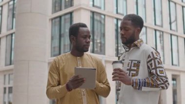 Gündüz vakti iş yerinde dışarıda dikilen, dijital tablete bakıp tartışan iki siyahi erkek iş arkadaşının orta boy fotoğrafı.
