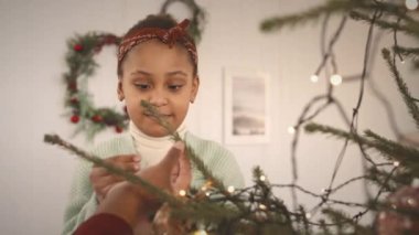 Güzel Afrikalı Amerikalı bir kızın beliyle ev yapımı Noel ağacını güzel oyuncaklarla süslerken annesi ona yardım ediyor.