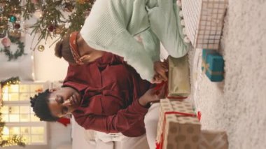 Dikey olarak Afrikalı Afrikalı Amerikalı küçük bir kız annesine Noel hediyesi olarak kırmızı kurdeleyle papyon bağlamasına yardım ederken, rahat dekore edilmiş bir dairede birlikte otururken.