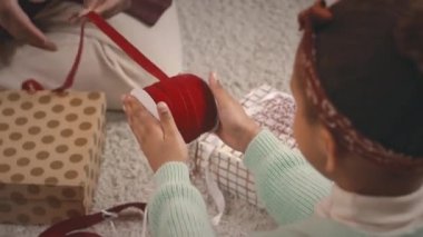 Yukarıdaki resimde küçük bir kız annesine Noel hediyelerini güzel kırmızı kurdeleli kutulara paketlemesi için yardım ederken dairede oturmuş elinde kurdele bobini tutarken.