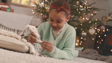 Mutlu Afrikalı 6 yaşındaki kız süslü Noel ağacının yanında halıda otururken pelüş tavşan oyuncağıyla oynuyor.