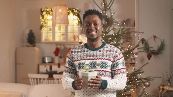 Xmasのために装飾された居心地の良いスタジオアパートに立っている手のクリスマスギフトボックスとカメラのためのクリスマスセーターの若い陽気な黒人のメディア肖像画 — ストック動画