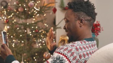 Yandan bakıldığında mutlu Afrikalı Amerikalı bir adamın Noel 'de uzakta olan akıllı telefonuyla ailesiyle görüntülü sohbetinin görüntüsü.