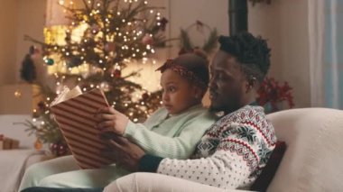 6 yaşındaki kızıyla birlikte Noel tatilinde rahat bir şekilde kanepede oturup kitap okuyan Afrikalı Amerikalı bir gencin orta boy fotoğrafı.