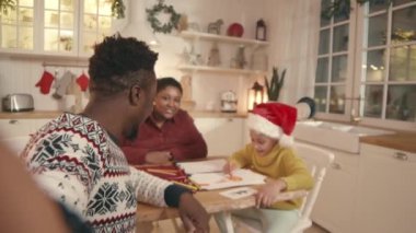 Noel süveteri giyen, ailesi ile birlikte mutfak masasında oturarak Noel videosu kaydeden neşeli, genç siyahi bir adamın el bilgisayarı.