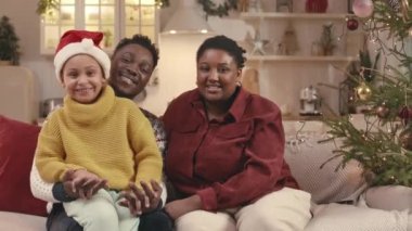 Noel için çam ağacı ve süslemelerle dekore edilmiş sıcak bir dairede koltukta otururken kameraya poz veren üç çocuklu mutlu Afrikalı Amerikalı ailenin orta boy portresi.