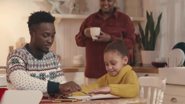 キッチンテーブルに座ってサンタに手紙を書く小さな娘にマシュマロとホットチョコレートでマグカップを提供する喜びの若い黒人女性のミディアムショット — ストック動画